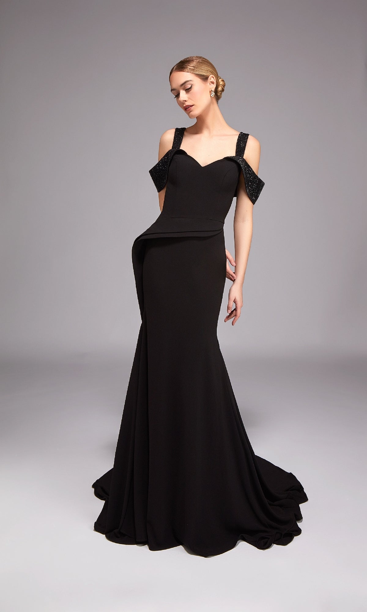 A woman poses elegantly against a plain gray background wearing the Alyce 27878 long formal black dress, featuring an off-the-shoulder style, fitted bodice, and flared skirt.